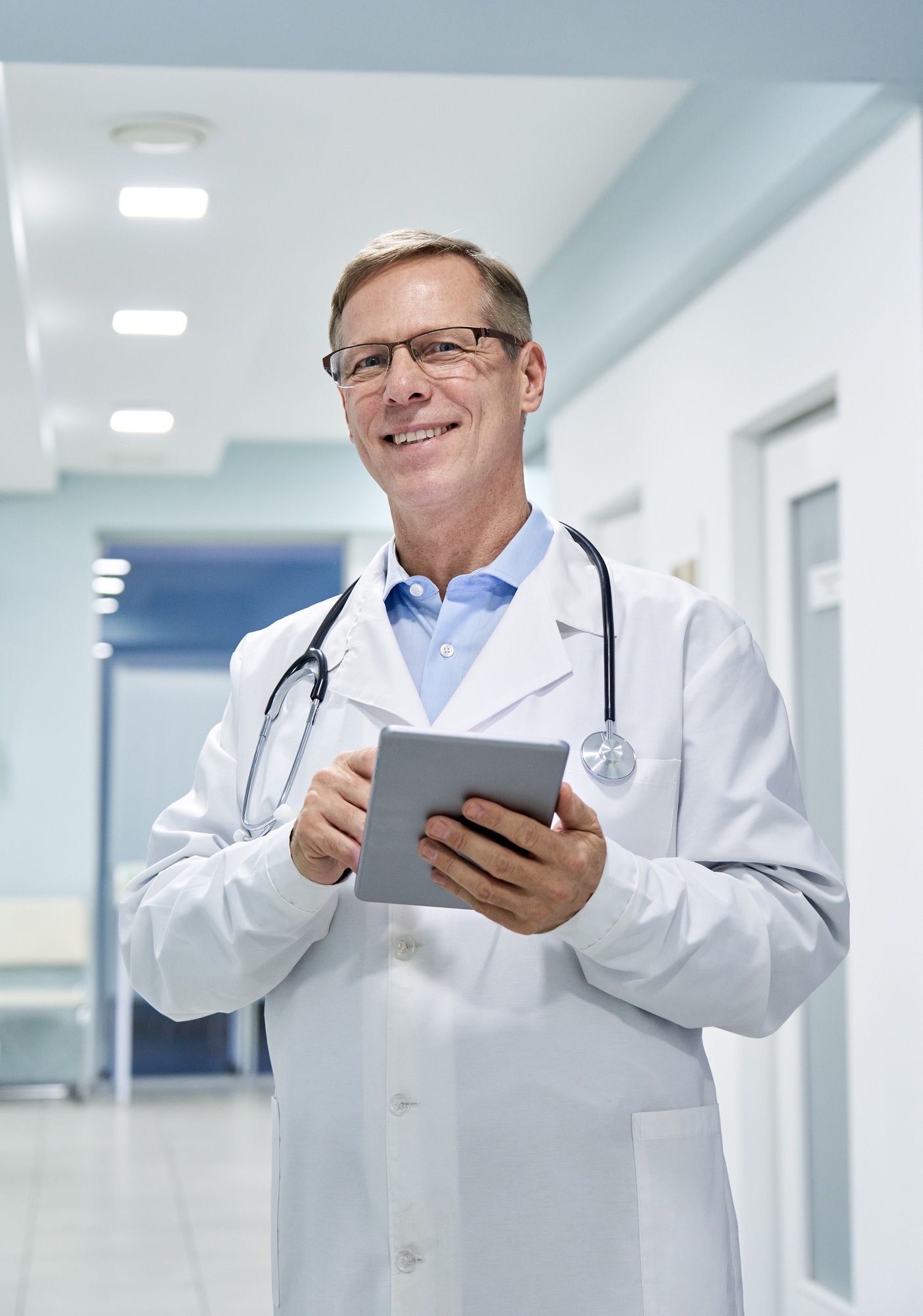 Medico anziano che tiene un tablet digitale guardando verso la fotocamera in ospedale.